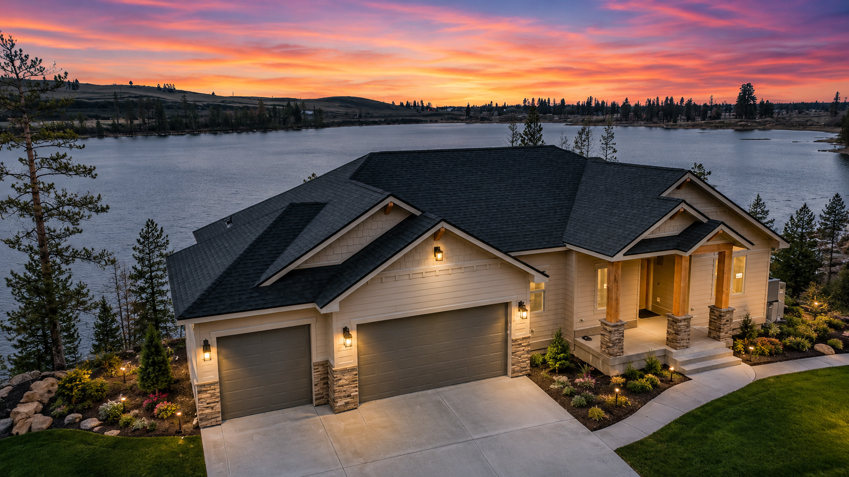 Aerial view of a craftsman-style custom home in North Idaho situated on a lakefront lot with a three-car garage and a vibrant sunset reflecting on the water.