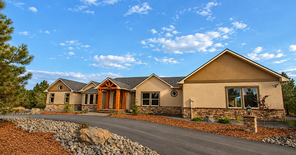 Exterior view of a custom single-story home featuring beige stucco, stone veneer accents, a wooden gabled entryway, and a paved curved driveway under a clear blue sky.