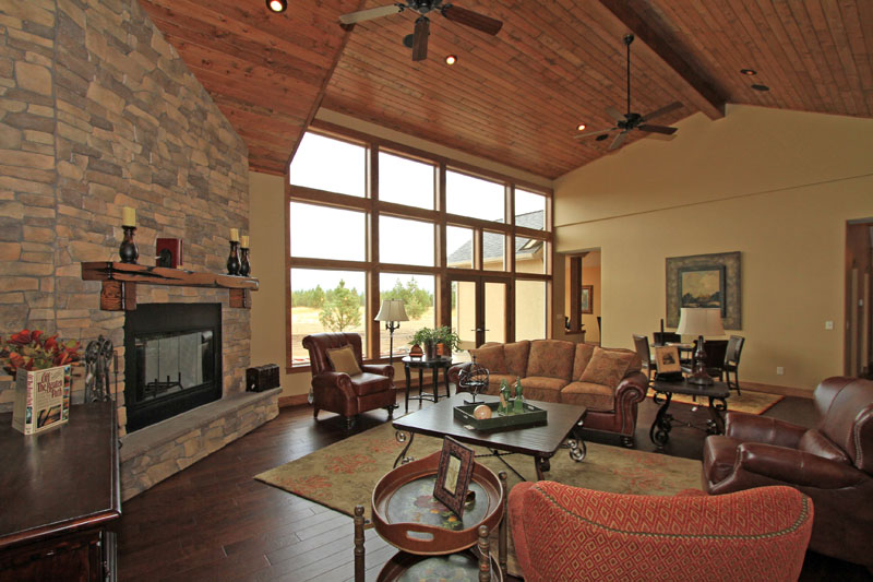 Spacious living room featuring a vaulted wood-plank ceiling, floor-to-ceiling windows, dark wood flooring, and a stone fireplace.