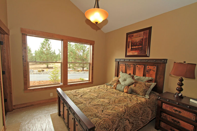 Warmly lit bedroom with a rustic wooden bed frame, copper panel accents, a matching nightstand, and large wood-trimmed windows.