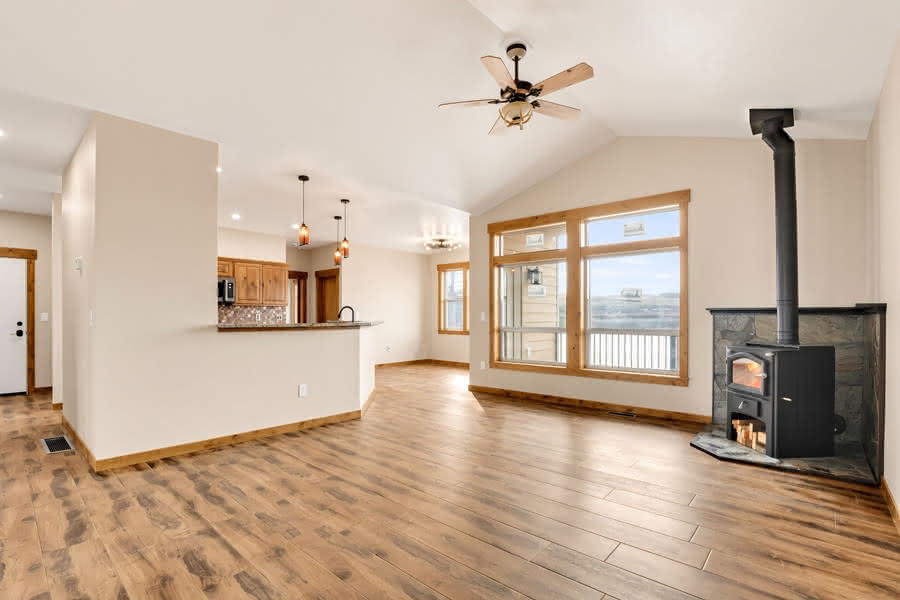 Spacious open-concept living area with wood-look flooring, vaulted ceiling, wood-burning stove, large windows, and an adjacent kitchen with breakfast bar.