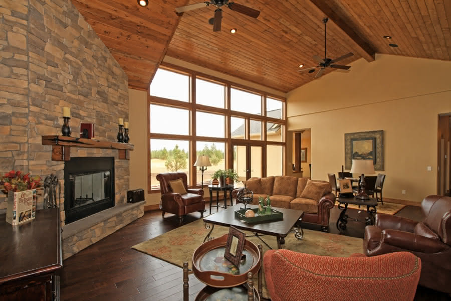 Rustic living room featuring a floor-to-ceiling stone fireplace, vaulted wood plank ceiling, dark hardwood floors, and a floor-to-ceiling wall of windows.