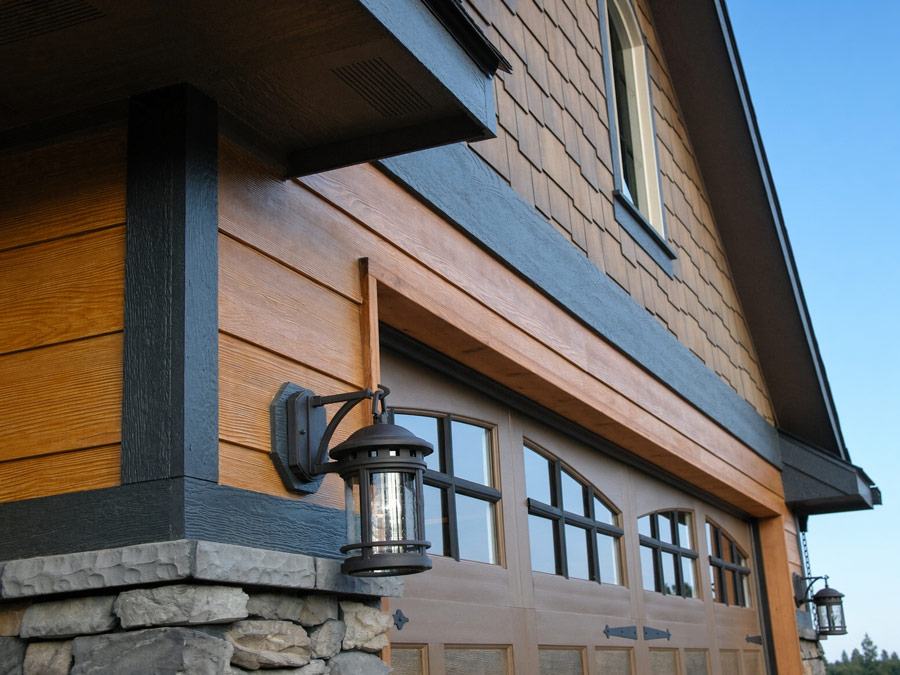 Low-angle view of a modern home exterior featuring warm wood lap siding, shake shingles, dark trim, stone accent masonry, and a black wall-mounted lantern light next to a carriage-style garage door.
